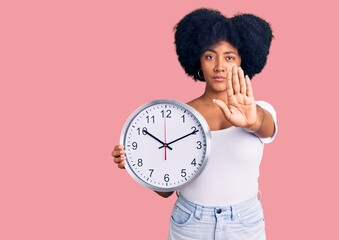 Sticker - Young african american girl holding big clock with open hand doing stop sign with serious and confident expression, defense gesture