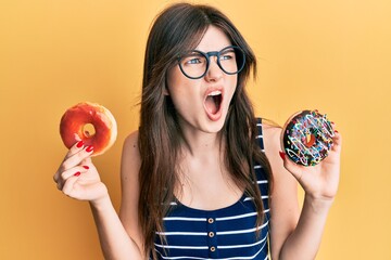 Sticker - Young beautiful caucasian girl holding tasty colorful doughnuts angry and mad screaming frustrated and furious, shouting with anger. rage and aggressive concept.