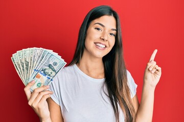 Sticker - Young brunette woman holding dollars smiling happy pointing with hand and finger to the side