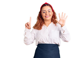 Young latin woman wearing waitress apron showing and pointing up with fingers number six while smiling confident and happy.