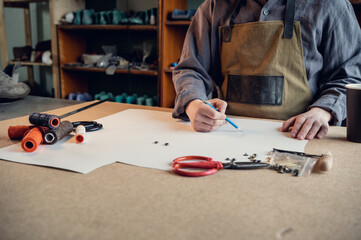 A young shoemaker makes a drawing for a pattern for leather shoes on a table in his workshop.