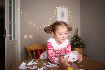 child cuts out snowflake at home in dining room at table on background of Christmas devoration. concept of new year kids creativity fun