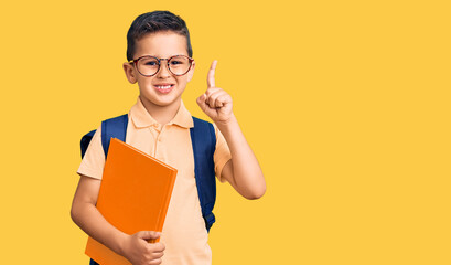 Wall Mural - Little cute boy kid wearing school bag and holding book surprised with an idea or question pointing finger with happy face, number one