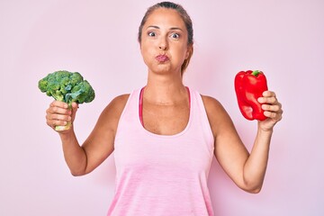 Poster - Middle age hispanic woman holding broccoli and red pepper puffing cheeks with funny face. mouth inflated with air, catching air.