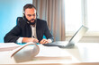 The man works remotely. Remote work of a bearded man. The boss communicates with subordinates via the Internet. A man in a business suit and headphones is sitting at a computer.