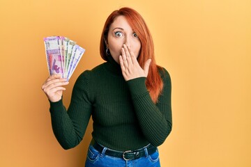 Poster - Beautiful redhead woman holding indian rupee banknotes covering mouth with hand, shocked and afraid for mistake. surprised expression