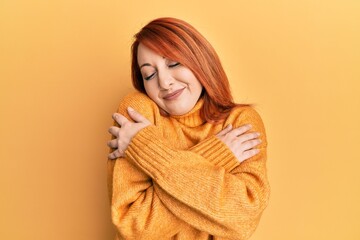 Poster - Beautiful redhead woman wearing casual winter sweater over yellow background hugging oneself happy and positive, smiling confident. self love and self care