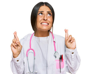 Beautiful hispanic woman wearing doctor uniform and stethoscope gesturing finger crossed smiling with hope and eyes closed. luck and superstitious concept.