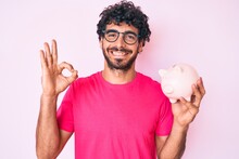 Handsome Young Man With Curly Hair And Bear Holding Piggy Bank Doing Ok Sign With Fingers, Smiling Friendly Gesturing Excellent Symbol