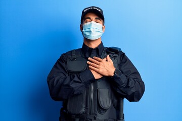 Poster - Young hispanic man wearing police uniform and medical mask smiling with hands on chest, eyes closed with grateful gesture on face. health concept.