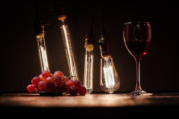 wine glass with wine and grapes nearby on a dark wooden table against the background of an edison lamp