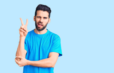 Young handsome man with beard wearing casual t-shirt smiling with happy face winking at the camera doing victory sign with fingers. number two.