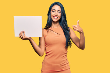 Wall Mural - Beautiful hispanic woman holding blank empty banner smiling happy and positive, thumb up doing excellent and approval sign