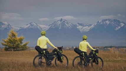 The man and woman travel on mixed terrain cycle touring with bikepacking. The two people journey with bicycle bags. Mountain snow capped.