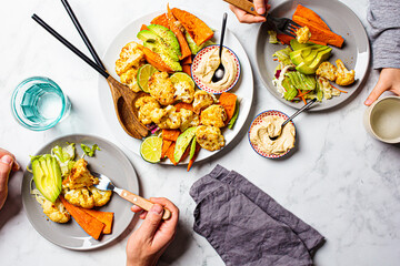 Canvas Print - Healthy food concept. Vegan dinner flat lay. People hands eating baked vegetables (sweet potato and cauliflower) with avocado and hummus, white marble background.