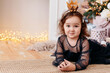 © Елена Лавринович - a little girl in a black dress lies on the floor against the background of a Christmas garland