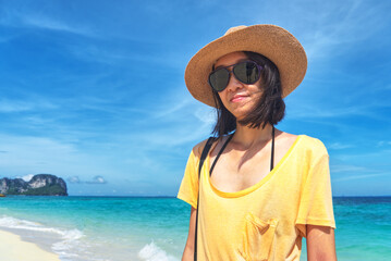 Portrait beautiful asian woman wear sunglasses and dress in yellow casual t-shirt and wear straw hat with little smile face standing in background turquoise sea and blue sky.