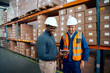 © StratfordProductions - Two male factory technician looking at digital tablet with stacked cardboard boxes at background