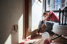 Side View Of Male Heating Engineer In Work Overalls Using Electric Drill While Laying Pipes On The Floor. Young Man Installing Heating System In Apartment. Concept Of Heating System Installation.