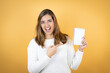 © Irene - Young caucasian woman over isolated yellow background smiling, surprised and pointing blank notebook in her hand