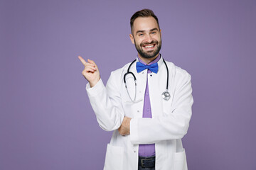 Wall Mural - Smiling young bearded doctor man wearing white medical gown pointing index finger up on mock up copy space isolated on violet background studio portrait. Healthcare personnel health medicine concept.