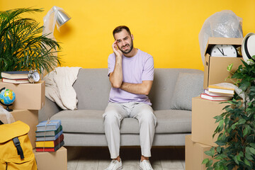 Wall Mural - Tired worried owner man put hand on head sits in living room on sofa at home household unpacking stuff in polythene indoor rents flat isolated on yellow wall. Relocation moving in apartment concept.