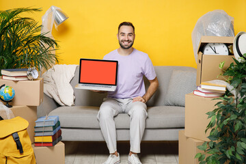 Wall Mural - Smiling owner man hold laptop pc computer with blank empty screen sits in room on sofa at home unpacking stuff indoor plant rents flat isolated on yellow wall. Relocation moving in apartment concept.