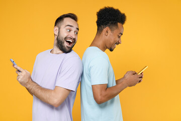 Wall Mural - Side view of excited young two friends european african american men 20s wearing violet blue casual t-shirts using mobile cell phone typing sms message isolated on yellow background studio portrait.