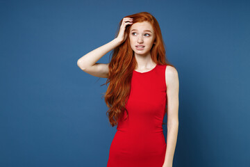 Wall Mural - Preoccupied puzzled pensive confused young redhead woman 20s wearing bright red elegant evening dress standing put hand on head looking aside isolated on blue color wall background studio portrait.