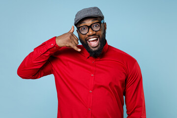 Excited young bearded african american man 20s wearing casual red shirt cap eyeglasses standing doing phone gesture like says call me back isolated on pastel blue color background studio portrait.