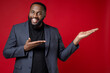 © ViDi Studio - Smiling cheerful funny young african american business man 20s wearing classic jacket suit standing pointing hands aside on mock up copy space isolated on bright red color background studio portrait.