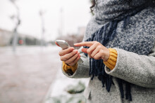 Young Woman Using Her Mobile Phone At A Snowy Winter Park. Closeup Of Female Checking News And Texting On Her Cellphone Outdoor During Cold Winter Season. Peoples' Gadgets Concept.