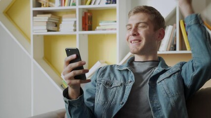 Smiling young man holds mobile phone while sitting on sofa at home and celebrating