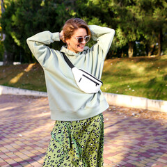 Wall Mural - Cheerful short-haired girl in brown glasses , green hoodie and green leopard skirt , smiles against urban park background.Attractive blonde in stylish clothes posing outdoor.
