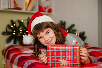 cute little happy smiling schoolgirl 8 years old wearing a santa hat and sitting at home on the bed holding a gift christmas and new year theme