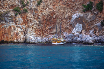 Wall Mural - Tourist yacht on the background of a steep cliff in the Mediterranean Sea in Alanya (Turkey). A boat trip along the rocky coast. Beautiful background with brown-orange mountain and turquoise water