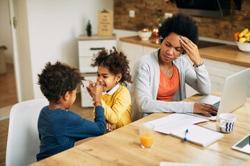 Wall Mural - Black working mother using laptop and feeling stressed out while kids are playing beside her.