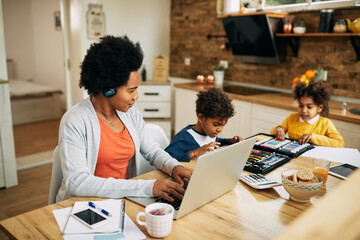 Wall Mural - Happy working black mother using laptop while her kids are coloring at home.