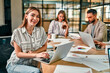 © Valerii Apetroaiei - Glad to be on the team. Beautiful cheerful woman with a laptop with a smile looks into the camera, sitting at an office table with her colleagues.
