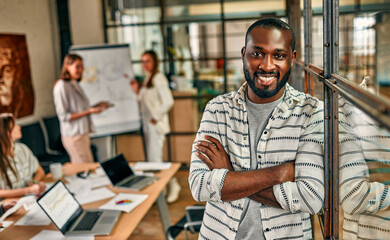 A young cheerful African American business man is standing with his arms crossed in a modern office. A successful hipster team in a coworking space. Freelancers.