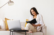 © foxyburrow - Beautiful brunette woman in white shirt sits on sofa in home office, holds pencil, black notepad in her hand.