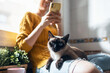 © nenetus - Pretty young woman with her cute cat using mobile phone while sitting on kitchen table at home.