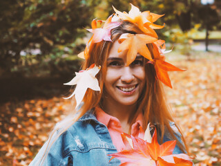 Wall Mural - Red-haired smiling girl with a head decorated with orange maple leaves. Autumn portrait outdoors