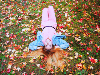 Wall Mural - A red-haired girl in a pink overalls lies on a meadow covered with autumn maple leaves