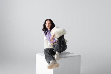 Wall Mural -  brunette young woman in stylish faux fur jacket posing on cube on white background