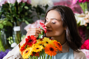 Smiling female florist with closed eyes smelling gerbera flower on blurred background