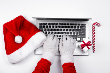 Santa Claus hands in medical latex gloves typing on laptop with Christmas gift, Santa's hat, gift box and Christmas candy can on white background.