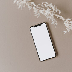 Blank screen mobile phone, white floral branch on neutral pastel beige background. Flat lay, top view minimalist lifestyle blog, website template. Copy space mockup.