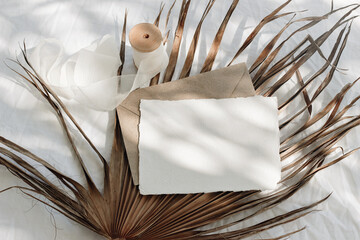 Wall Mural - Tropical stationery still life. Closeup of blank card mock-up and craft envelope in sunlight. Dry palm leaf , white linen table cloth background. Summer vacation concept. Boho design. Flat lay, top.