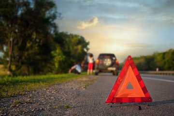 Close-up of triangular road warning sign with couple standing in front of broken car. at sunset